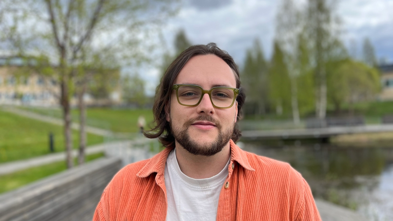 A man with long hair and glasses standing in front of a body of water.