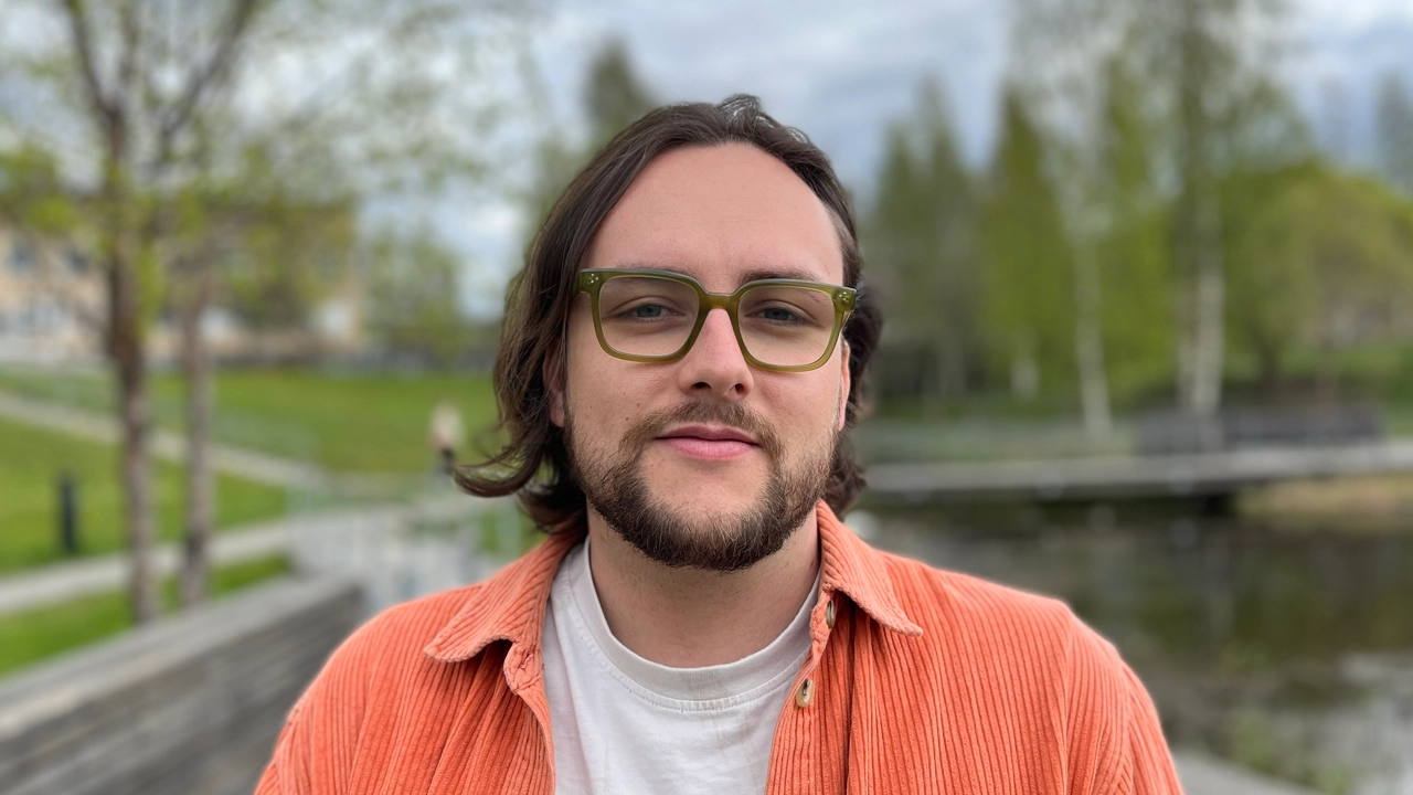 A man with long hair and glasses standing in front of a body of water.