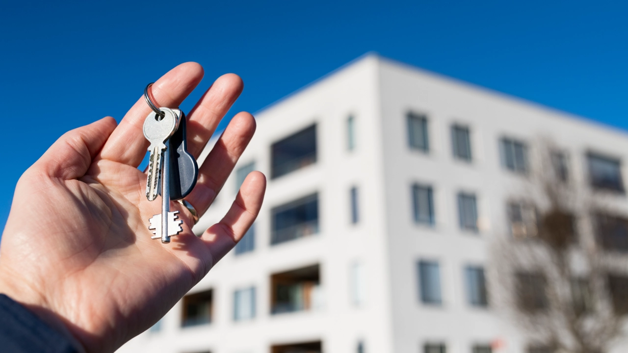 Hand med nycklar till en lägenhet. Lägenhetshus i bakgrunden.