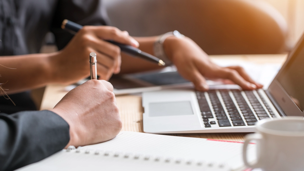 The hands of two people collaborating, in front of a laptop standing on a desk.