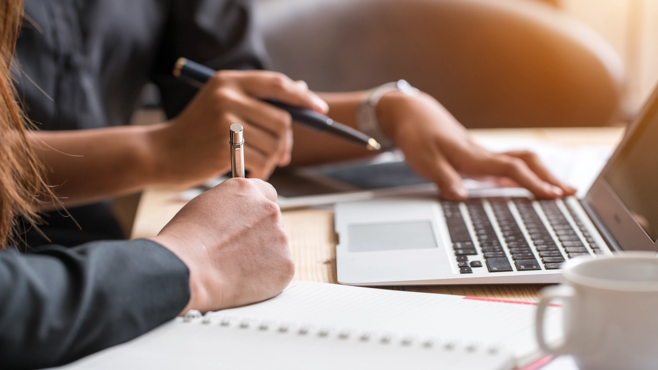 Närbild på två personers händer med dator, pennor och papper för att illustrera samarbete eller grupparbete.