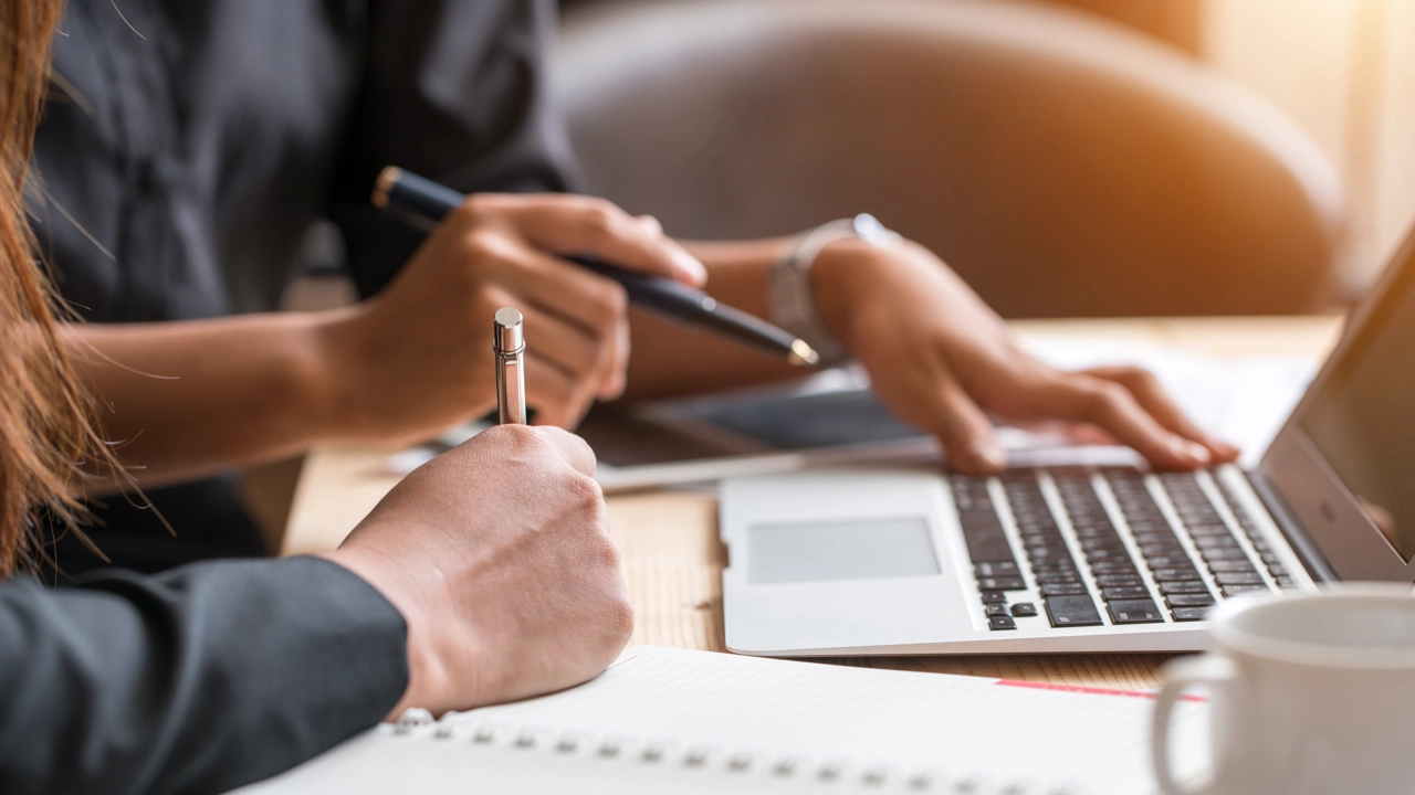 Närbild på två personers händer med dator, pennor och papper för att illustrera samarbete eller grupparbete.