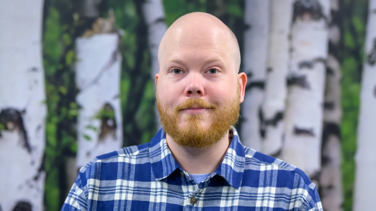 A man in a blue plaid shirt with a beard in front of a birch wallpaper background