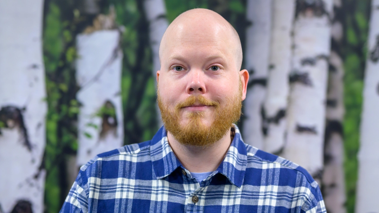 En man i en blå rutig skjorta med skägg framför en björktapetbakgrund