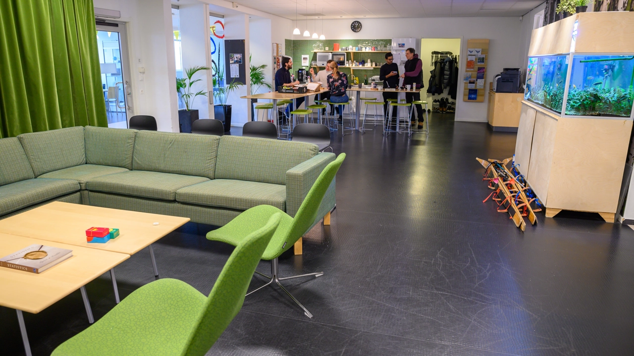 Vardagsrum och kök fyllt med möbler och ett akvarium. Några personer har samlats vid de bortre borden i köksdelen.