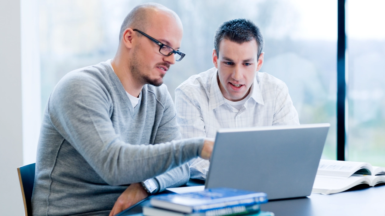 Two men at a table looking at a laptop screen.