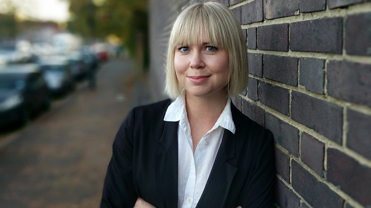 Porträttbild på Jennifer Eriksson, mörk tegelvägg och gata i bakgrunden