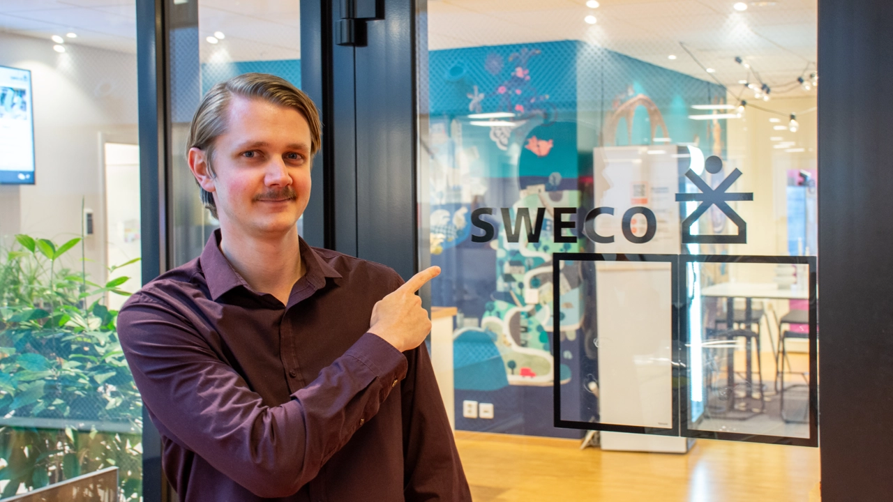 Bild på Jesper Lagerström vid Swecos entré, pekar på företagsskylten