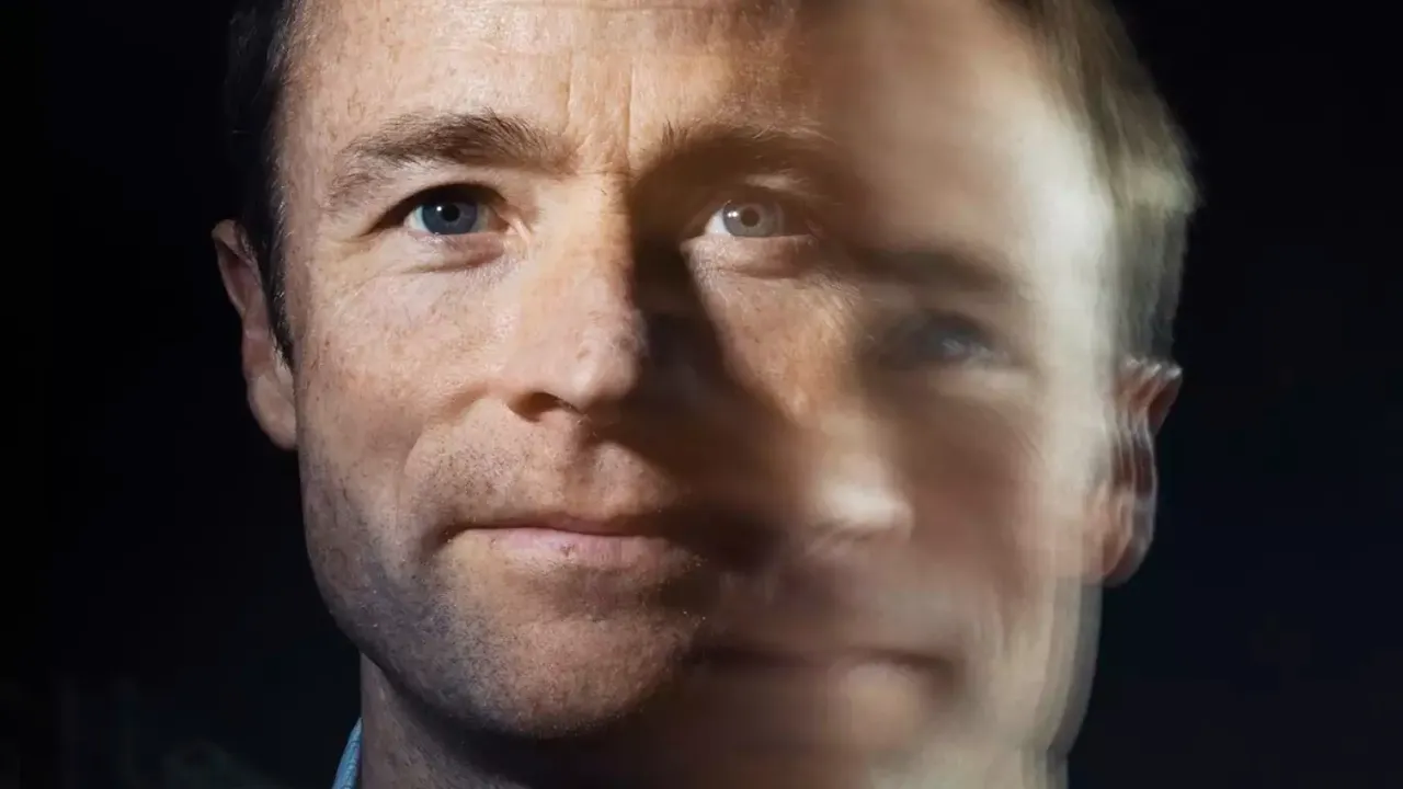 A man with short brown hair looking at the camera. The image is double exposed so it appears as though there is a blurry second image of the man to the right and slightly below the sharp image. 