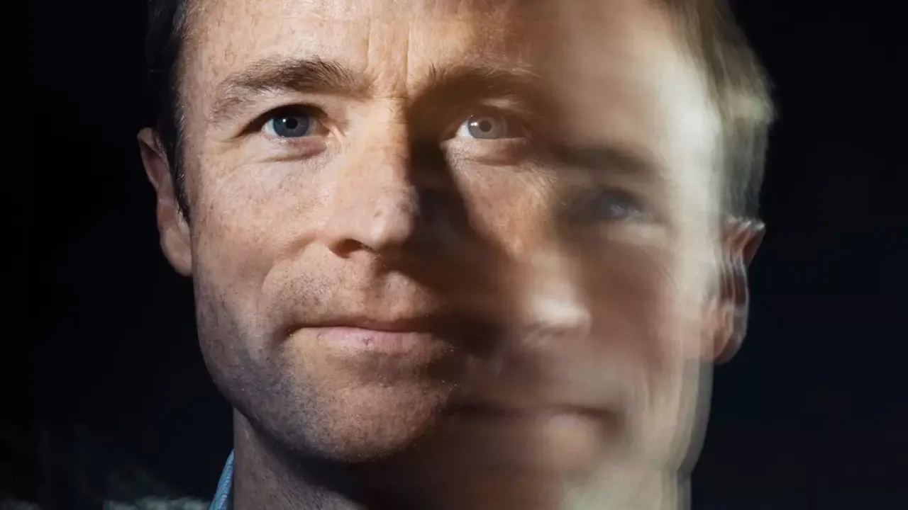 A man with short brown hair looking at the camera. The image is double exposed so it appears as though there is a blurry second image of the man to the right and slightly below the sharp image. 