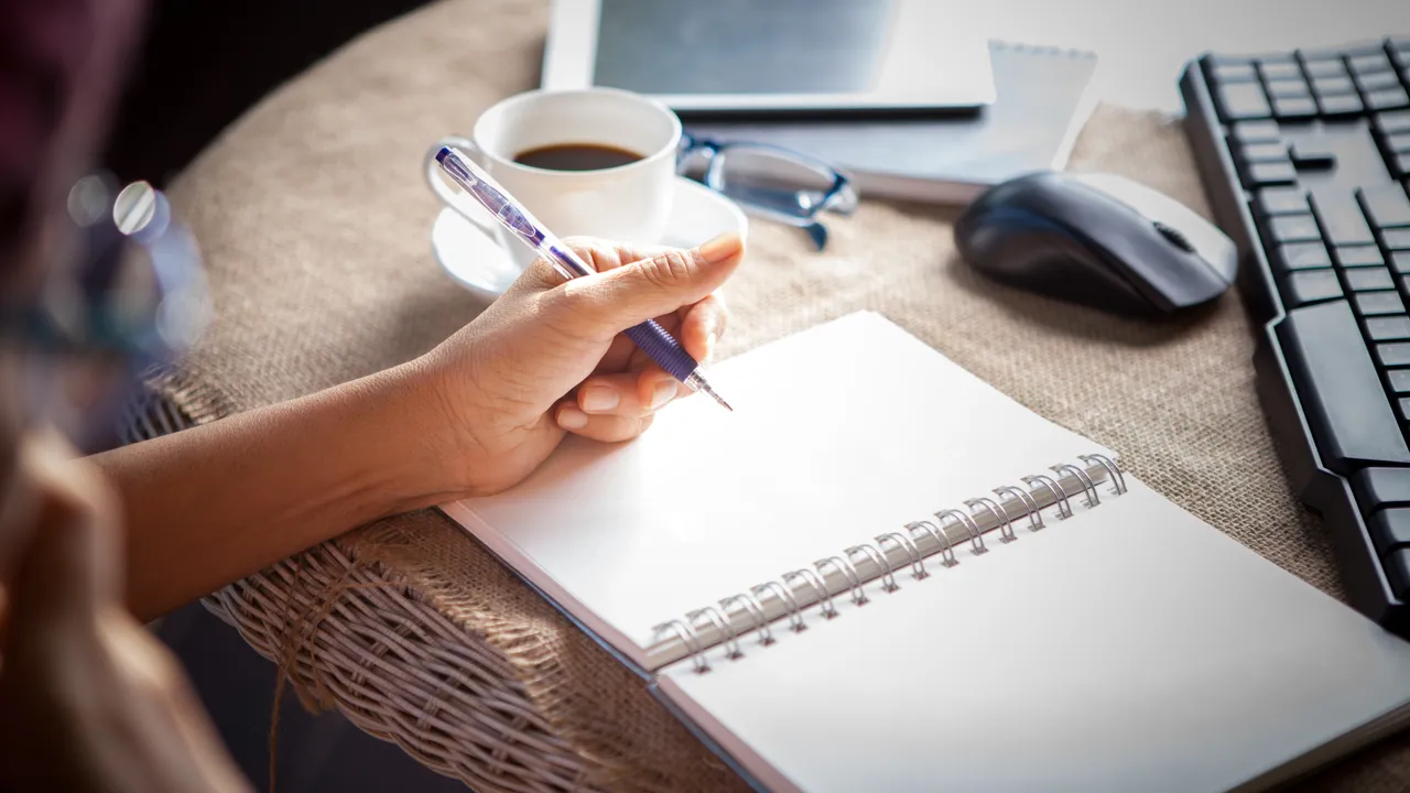 Person sitter med penna, block och kaffekopp framför dator. 