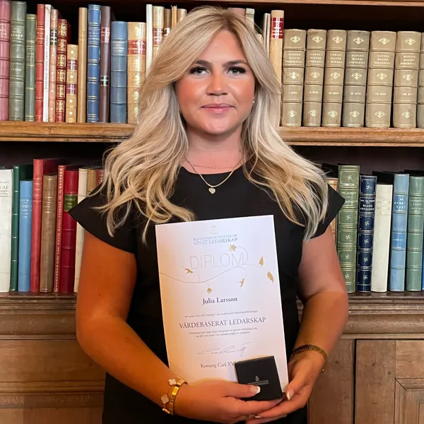 Bild på Julia Larsson stående framför bokhylla, håller upp ett diplom