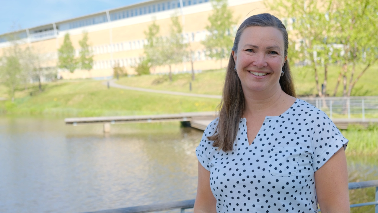 Karin Åström is standing in front of the Social science building by the pond. 