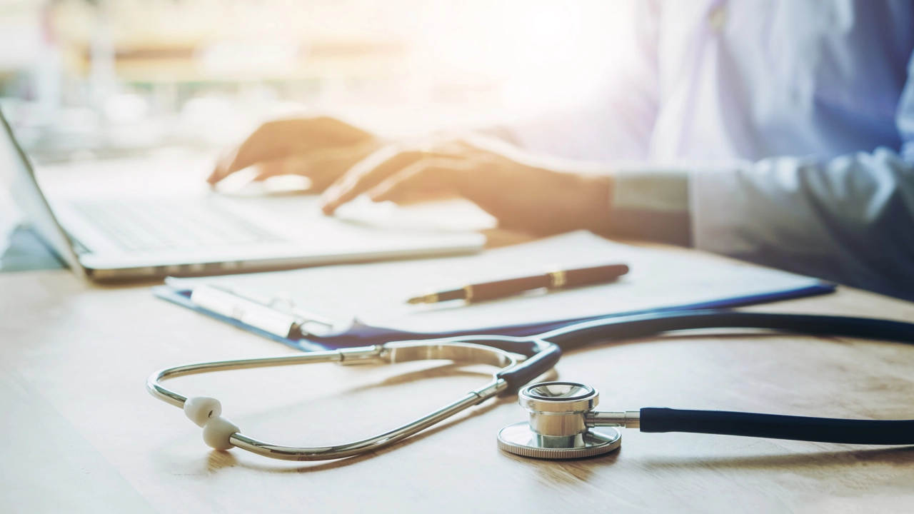 Ett stetoskop ligger på ett bord medan en läkare utför administrativa uppgifter på en dator.