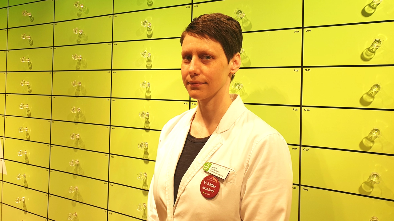 Bild på Livia Karlström stående mot en vägg med gula lådor på ett apotek