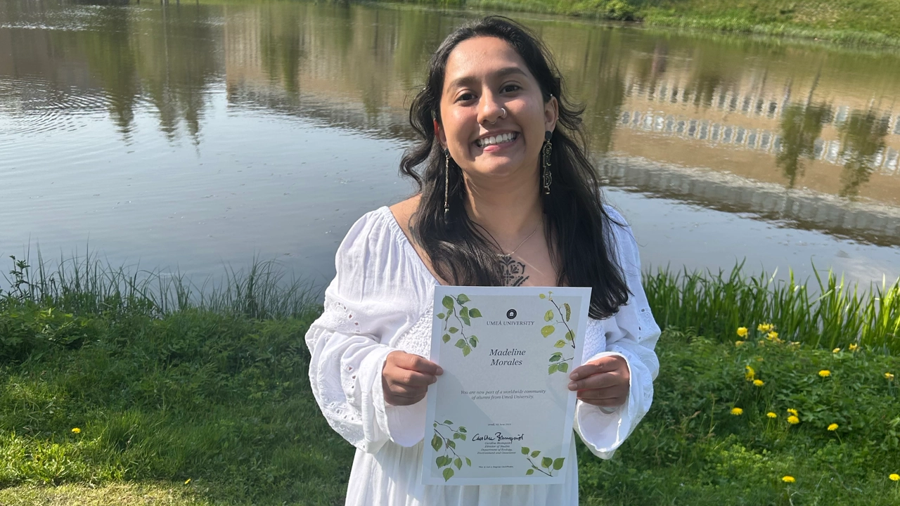 Photo of Madeline Morales holding her master's diploma, the campus pond in the background