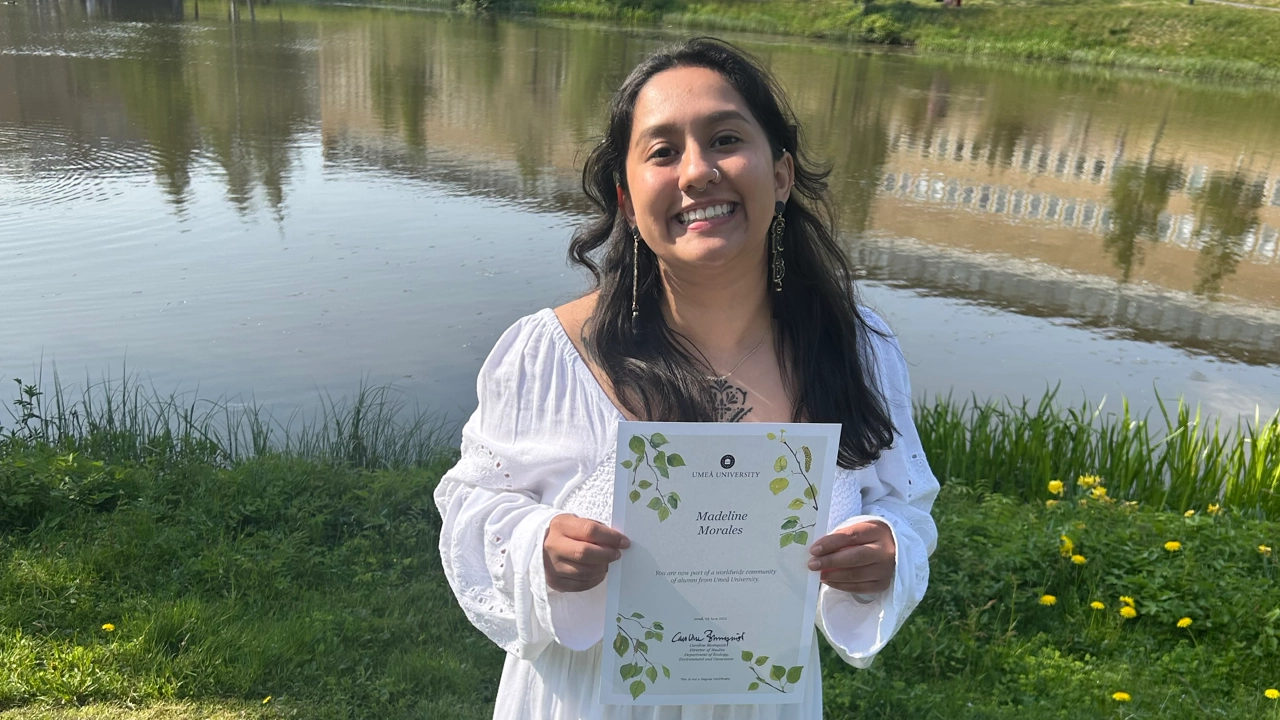 Foto av Madeline Morales med sitt masterdiplom i händerna, i bakgrunden campusdammen