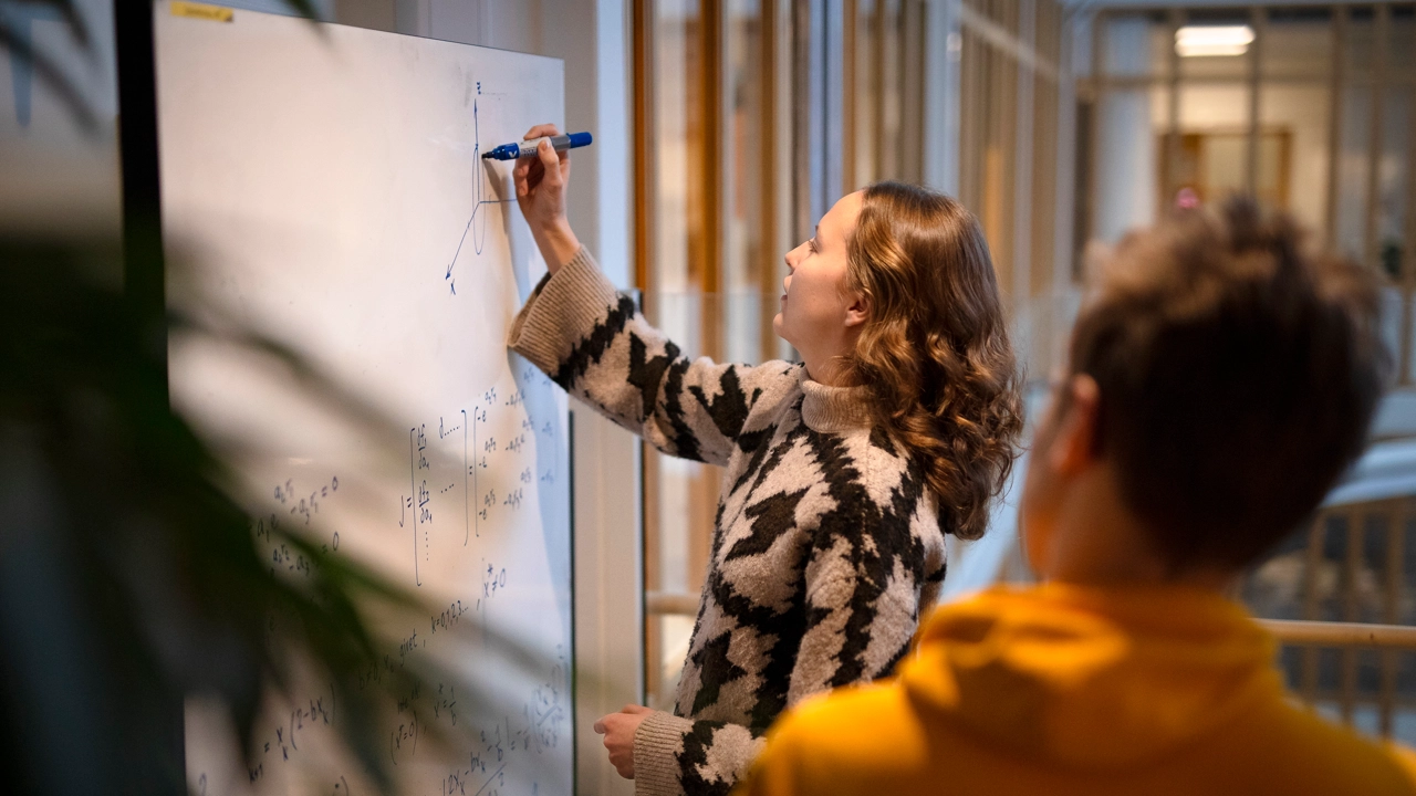 Kvinna skriver formler på whiteboard medan en man tittar på. 