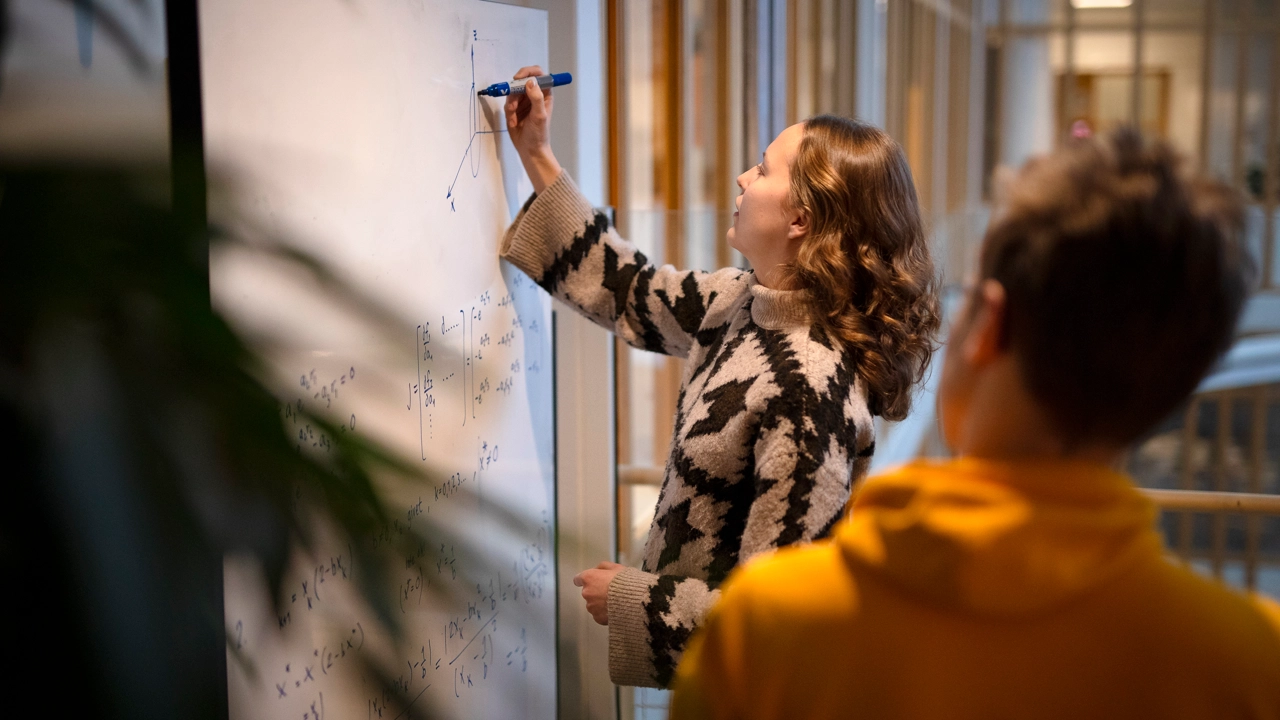 A woman is writing formulas on a whiteboard while a man is watching.
