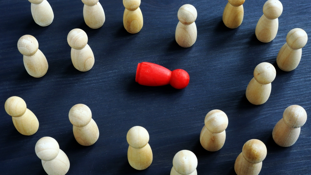 Små spelpjäser i trä står runt en liggande rödfärgad spelpjäs. För att illustrera mobbning eller trakasserier.