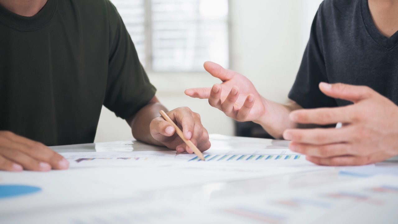 Detalj på två personer som sitter vid ett bord med papper och pratar med varandra.