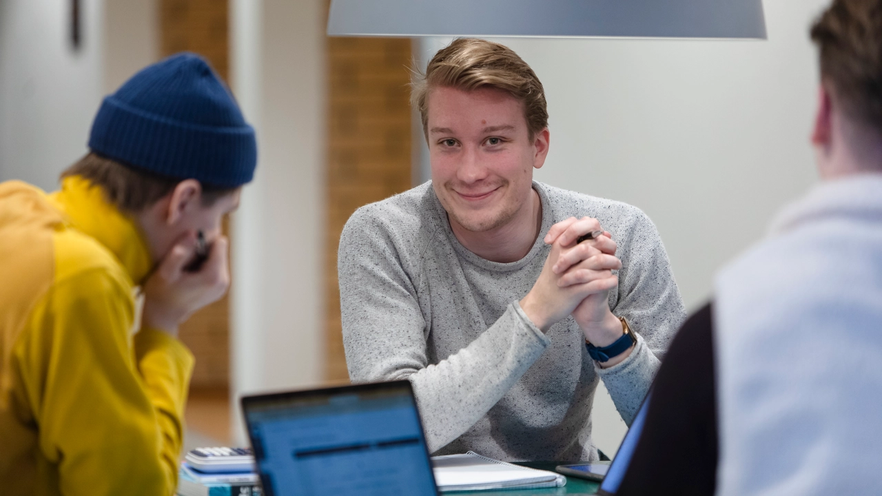 En leende kille tittar rakt in i kameran. Framför honom sitter studiekamrater vid en studieplats på campus.