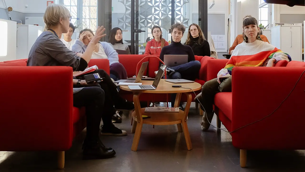 Tio personer sitter samlade runt ett bord. En talar och gestikulerar medan de andra lyssnar koncentrerat. I högra kanten syns baksidan av en monitor.