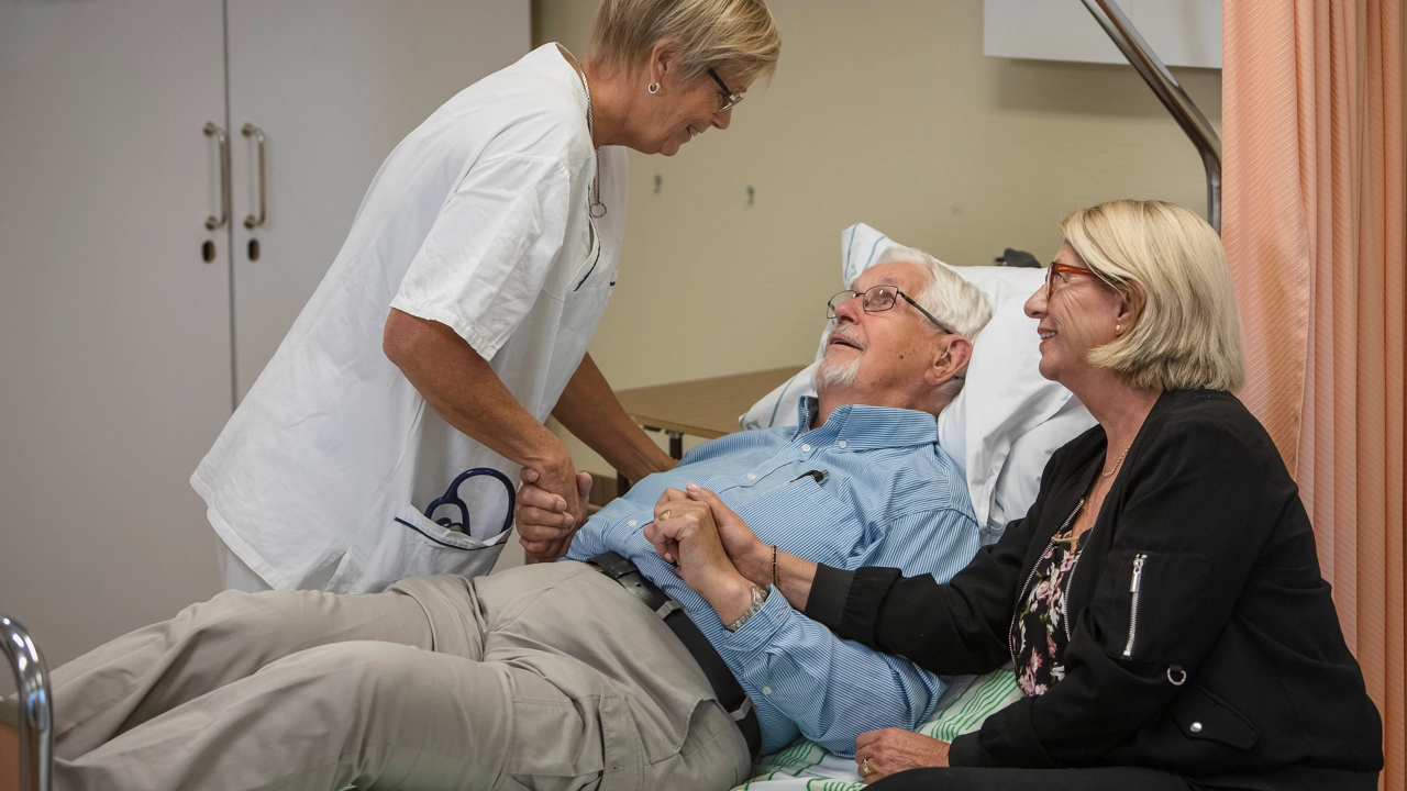 En sjuksköterska tar en liggande patient i hand. Hans anhöriga sitter på en stol och håller i den andra handen.