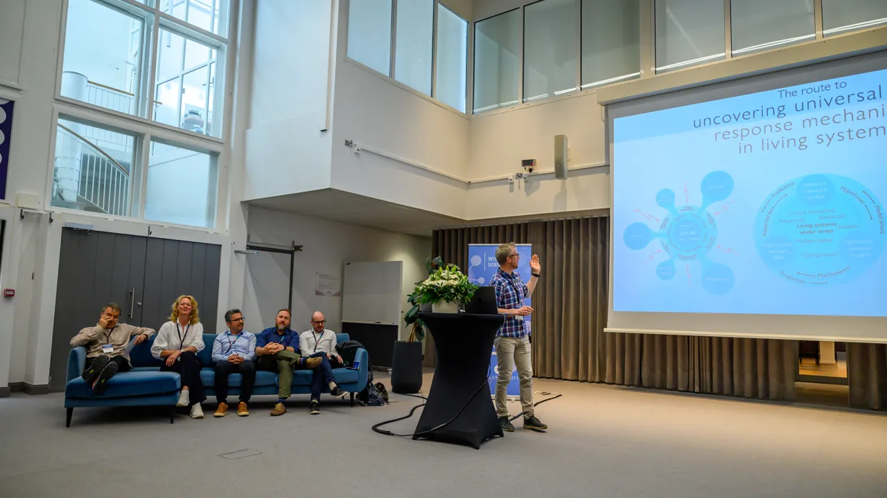 Five people sit on a blue sofa and one stands speaking next to a large screen.