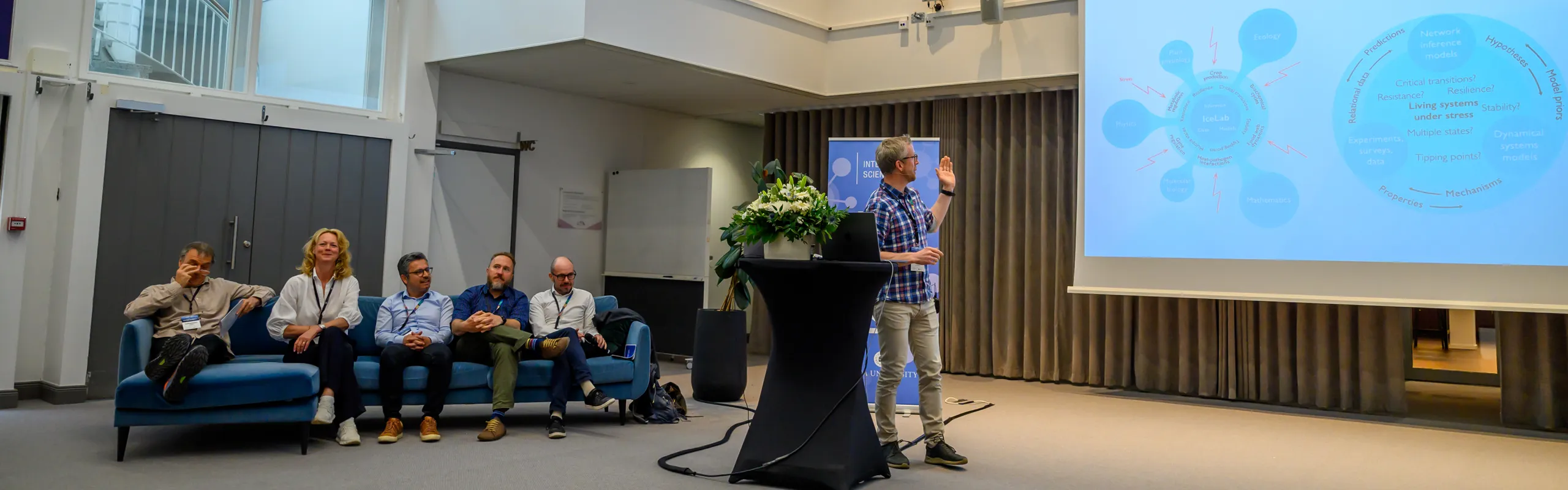Five people sit on a blue sofa and one stands speaking next to a large screen.
