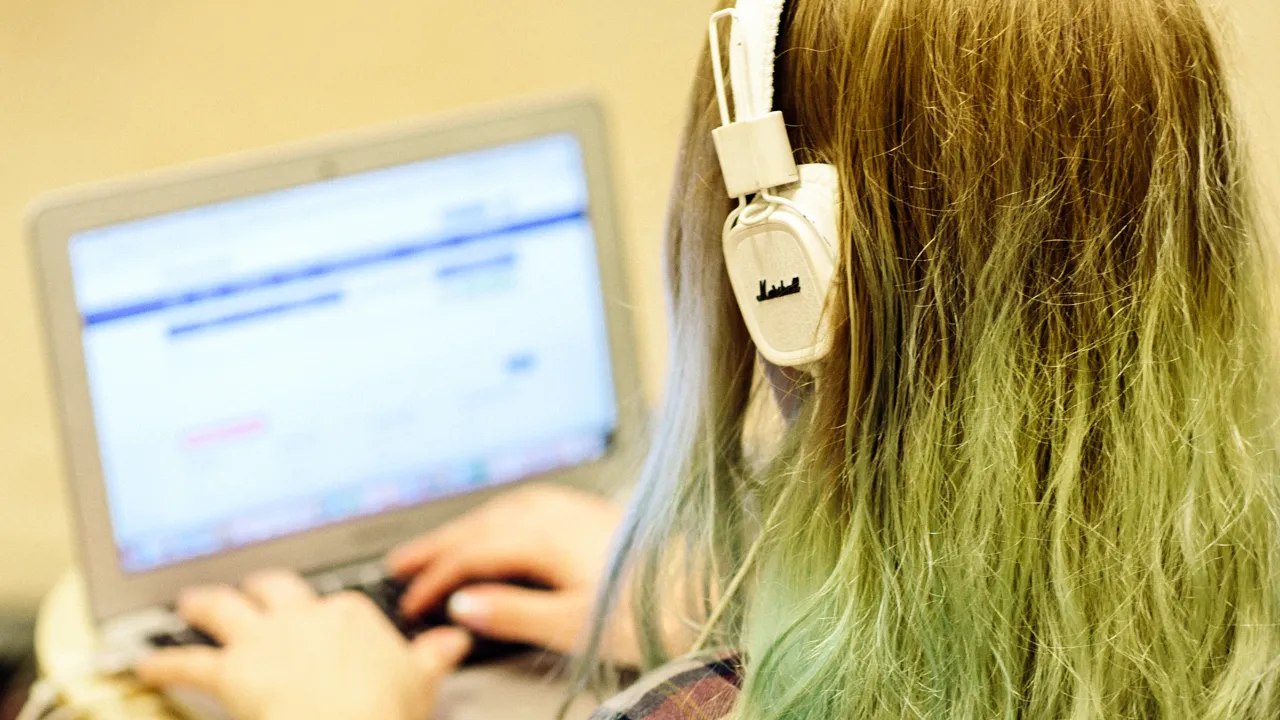 A person with long hair and headphones is sitting with their back to the camera, working on a laptop. 
