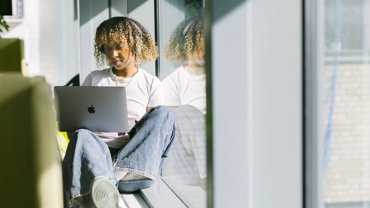 En student sitter i en solig fönstersmyg och läser på en laptop. 