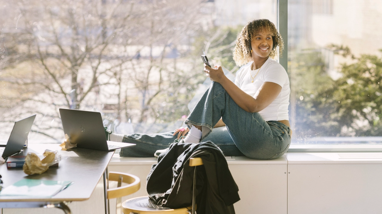 Student sitter i fönster med en mobiltelefon och dator.