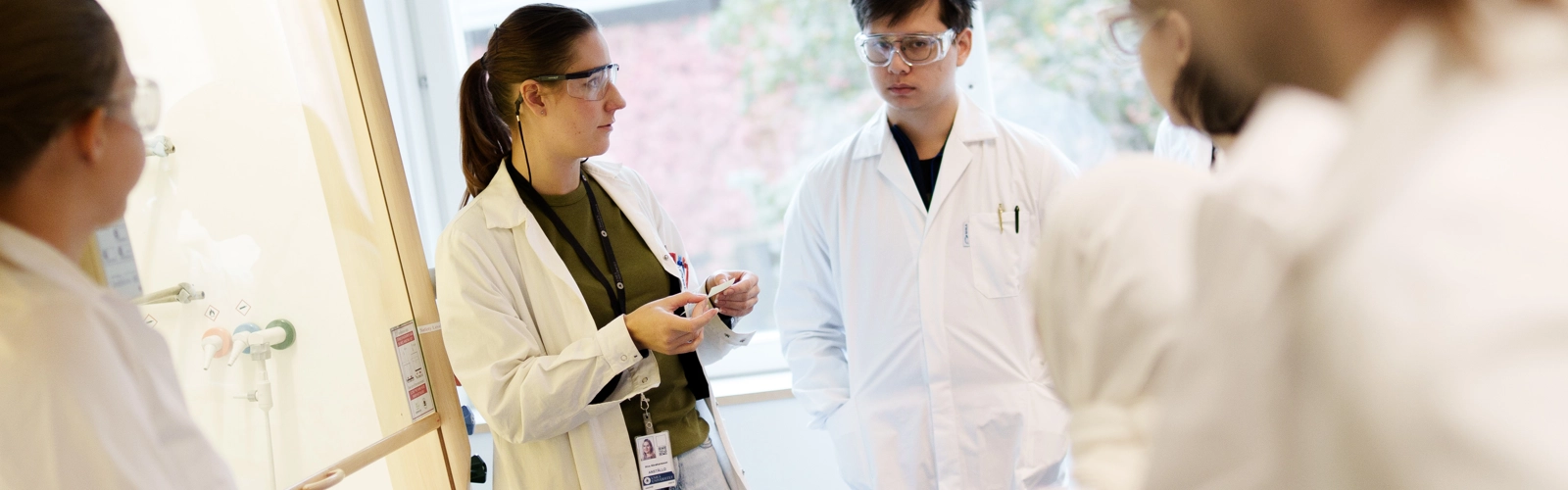 Bild på lärare och en grupp studenter i labbrockar under genomgång.