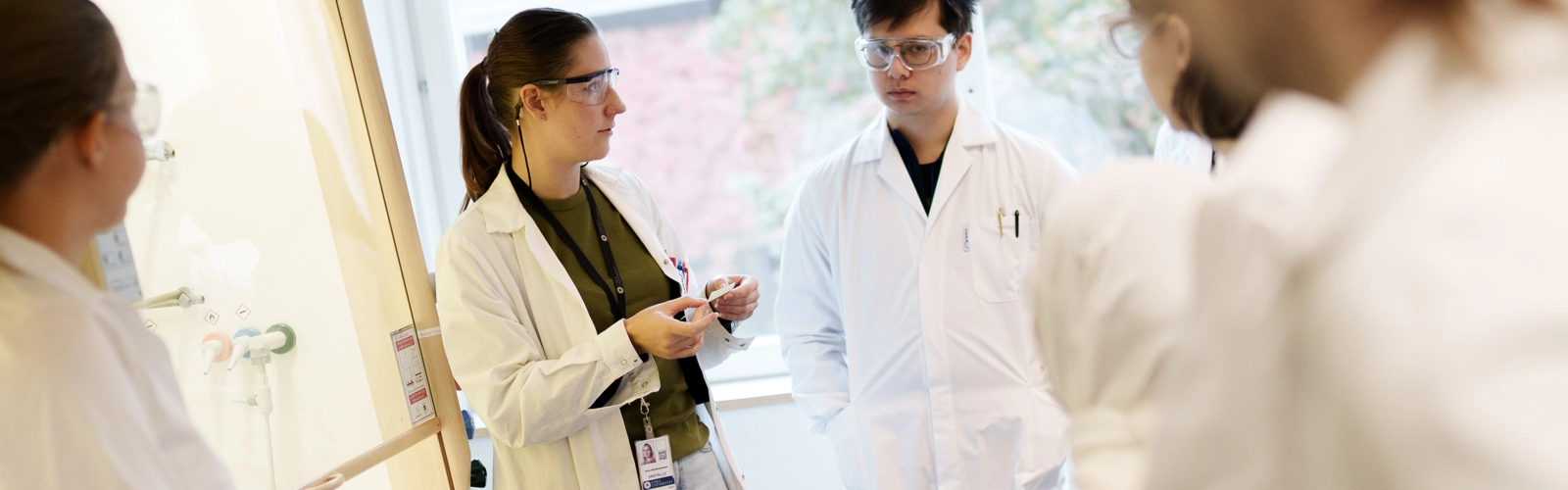 Bild på lärare och en grupp studenter i labbrockar under genomgång.