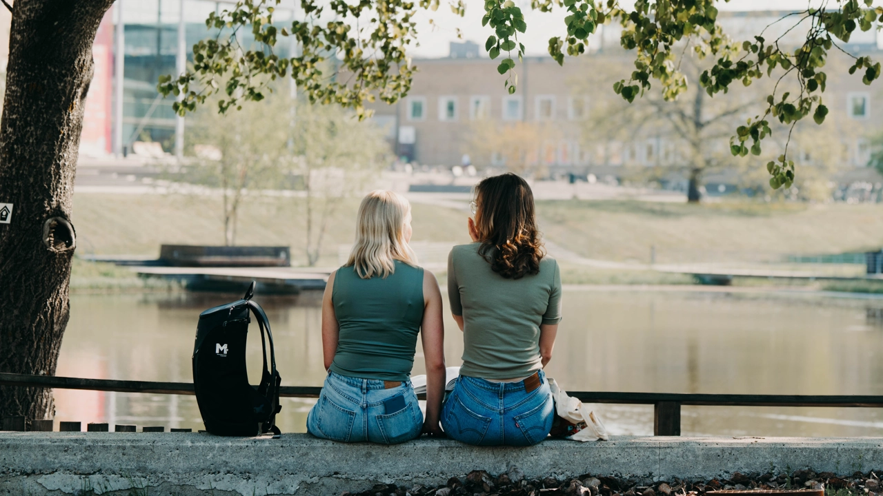 Två studenter sitter vid campusdammen med ryggen mot kameran.