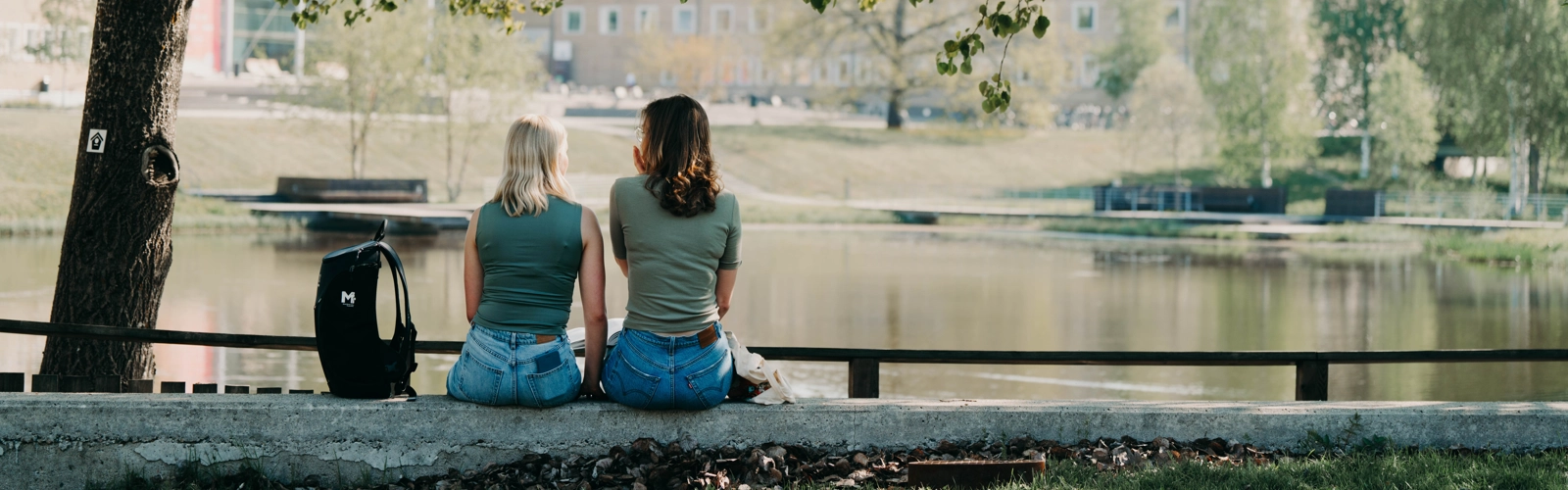 Två kvinnor sitter vid campusdammen. Ryggen mot kameran. Under ett träd.
