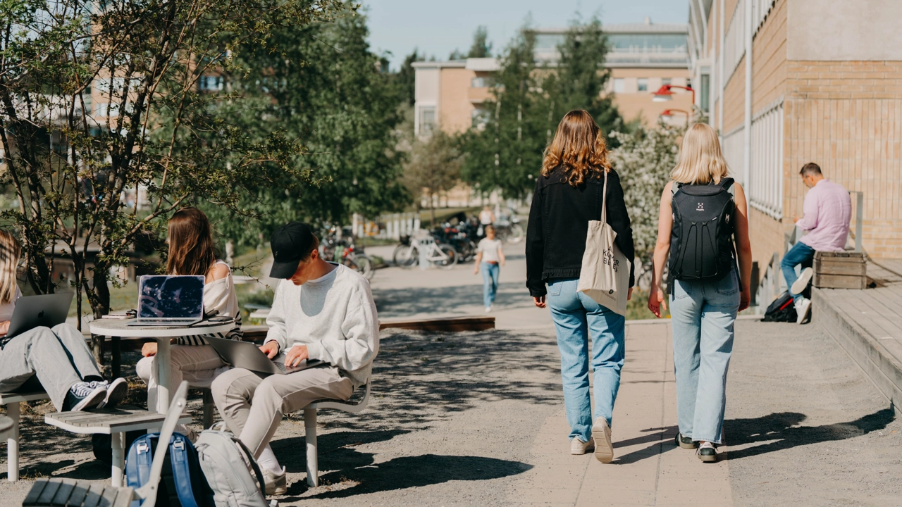 Två kvinnor går på Umeå campus. Det sitter personer vid bord runt om. 