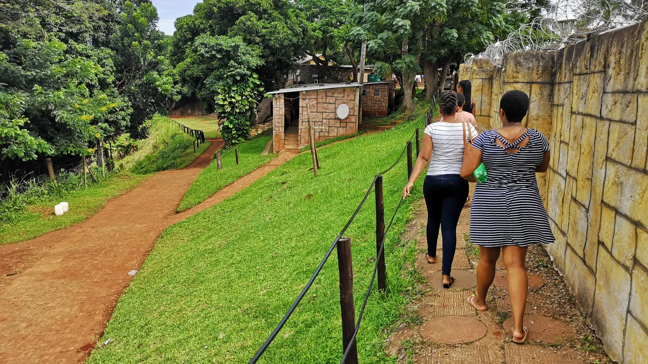 Tre kvinnor går längs en stig med en mur och ett staket på vardera sida i Sydafrika.