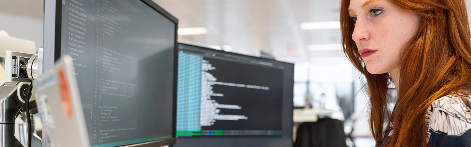 Kvinna med långt rött hår håller koll på dataskärmar och information.