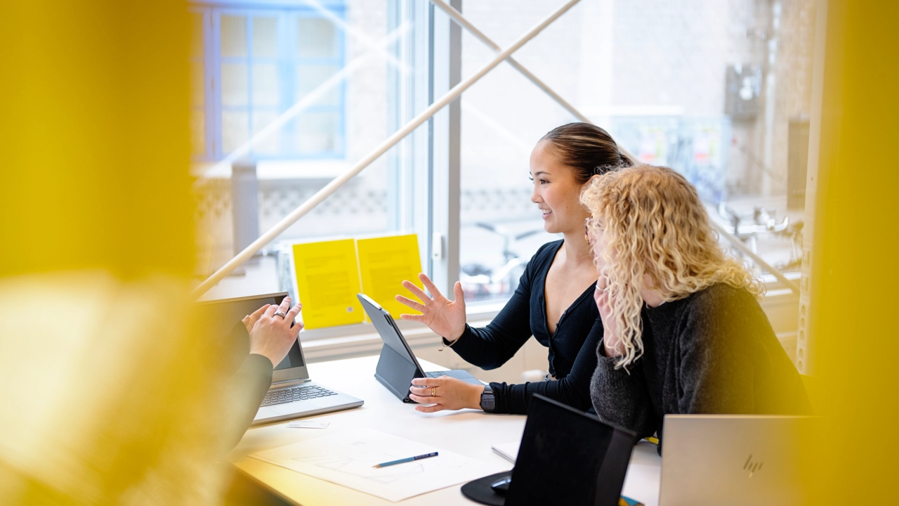 Två kvinnor pratar med en tredje person som skymtar på andra sidan ett skrivbord. 