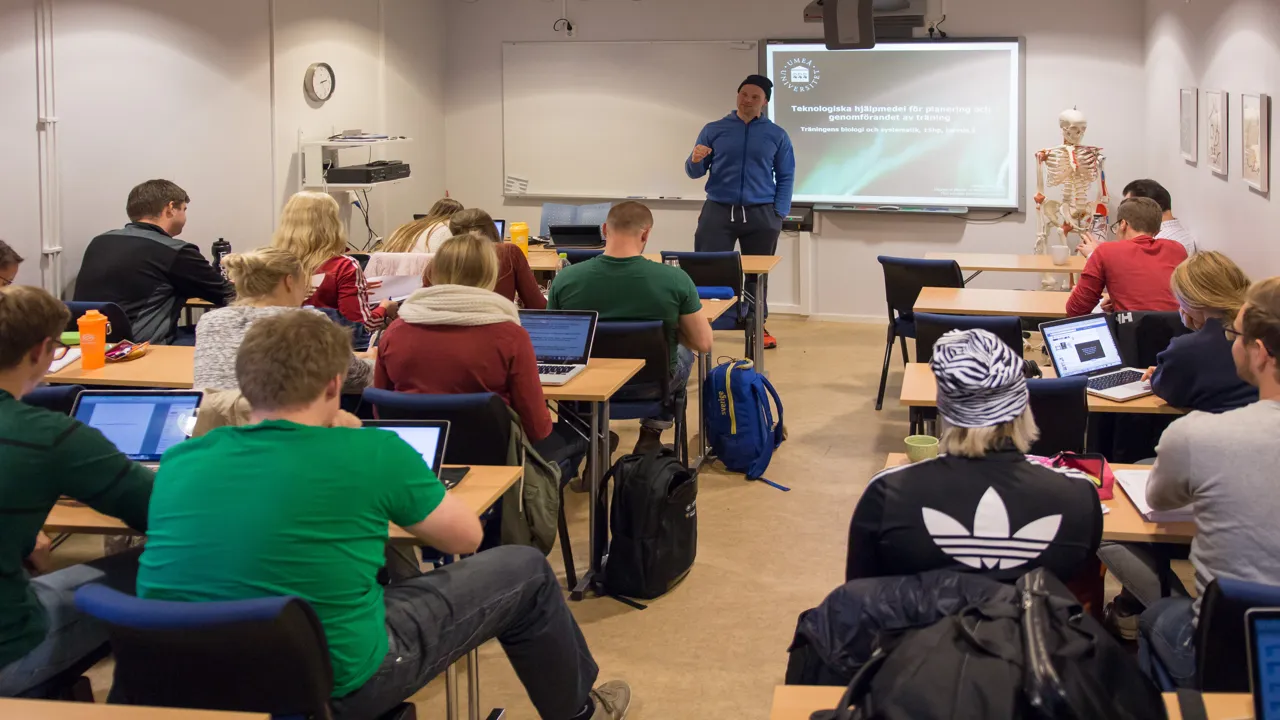 Ett klassrum med studenter sittande med ryggen mot kameran och en manlig lärare som står längst fram.