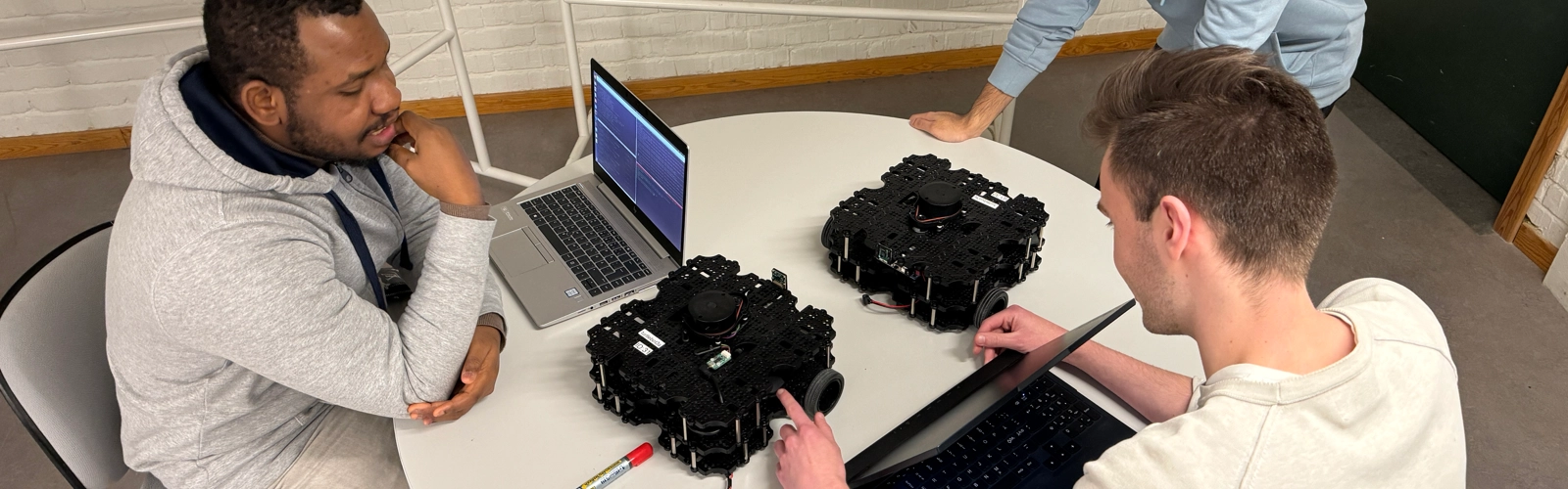 Tre studenter sitter ned kring ett runt bord. De har datorer och två robotar på hjul. De diskuterar, bakom dem finns det en tavla med matematiska beräkningar.