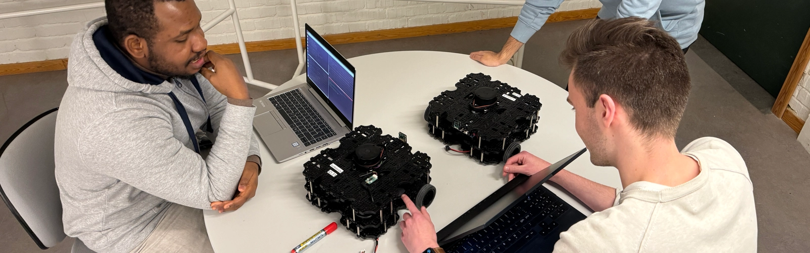 Tre studenter sitter ned kring ett runt bord. De har datorer och två robotar på hjul. De diskuterar, bakom dem finns det en tavla med matematiska beräkningar.