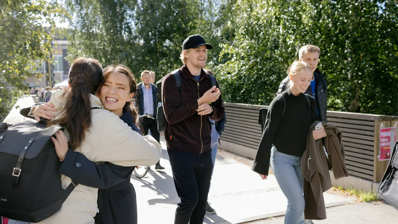 Två personer kramar varandra utomhus. Tre andra personer går i bakgrunden.