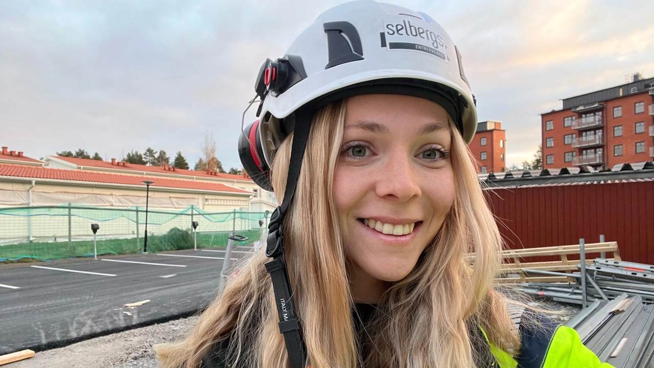 Porträttbild på Wilma Edin i vit bygghjällm, husbygge i bakgrunden