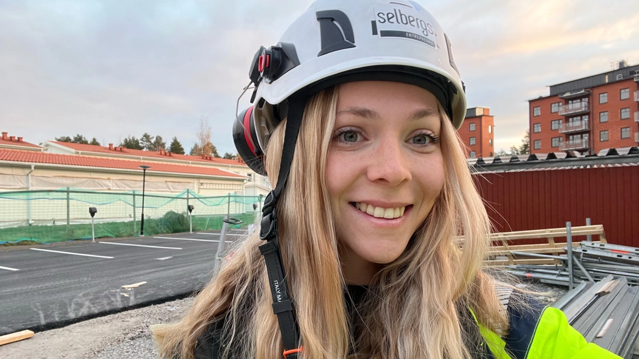 Porträttbild på Wilma Edin i vit bygghjällm, husbygge i bakgrunden