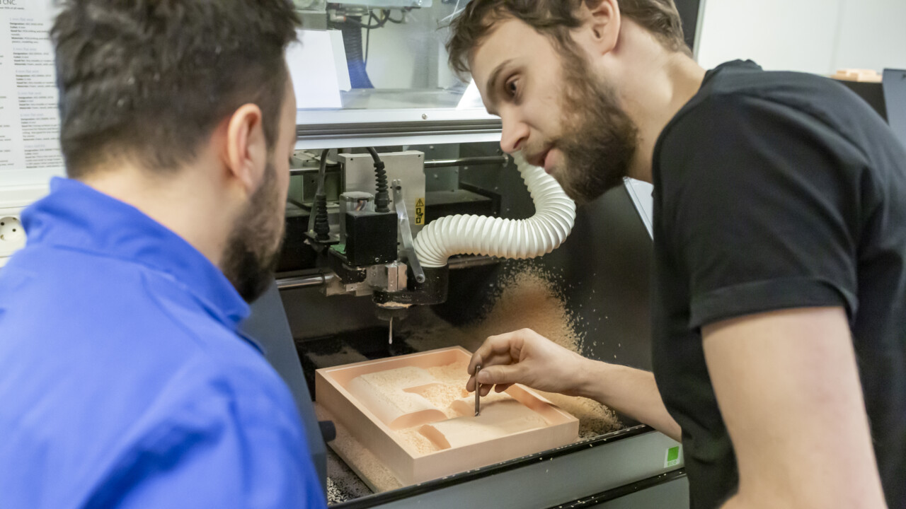 Två personer står framåtlutade över en verkstadsmaskin (3D-fräs). Vi ser dem bakifrån i närbild. Den ene tittar på den andre och håller ett frässtål i högra handen. Han liksom pekar med frässtålet och man förstår av situationen att han förklarar någo för den andra personen.