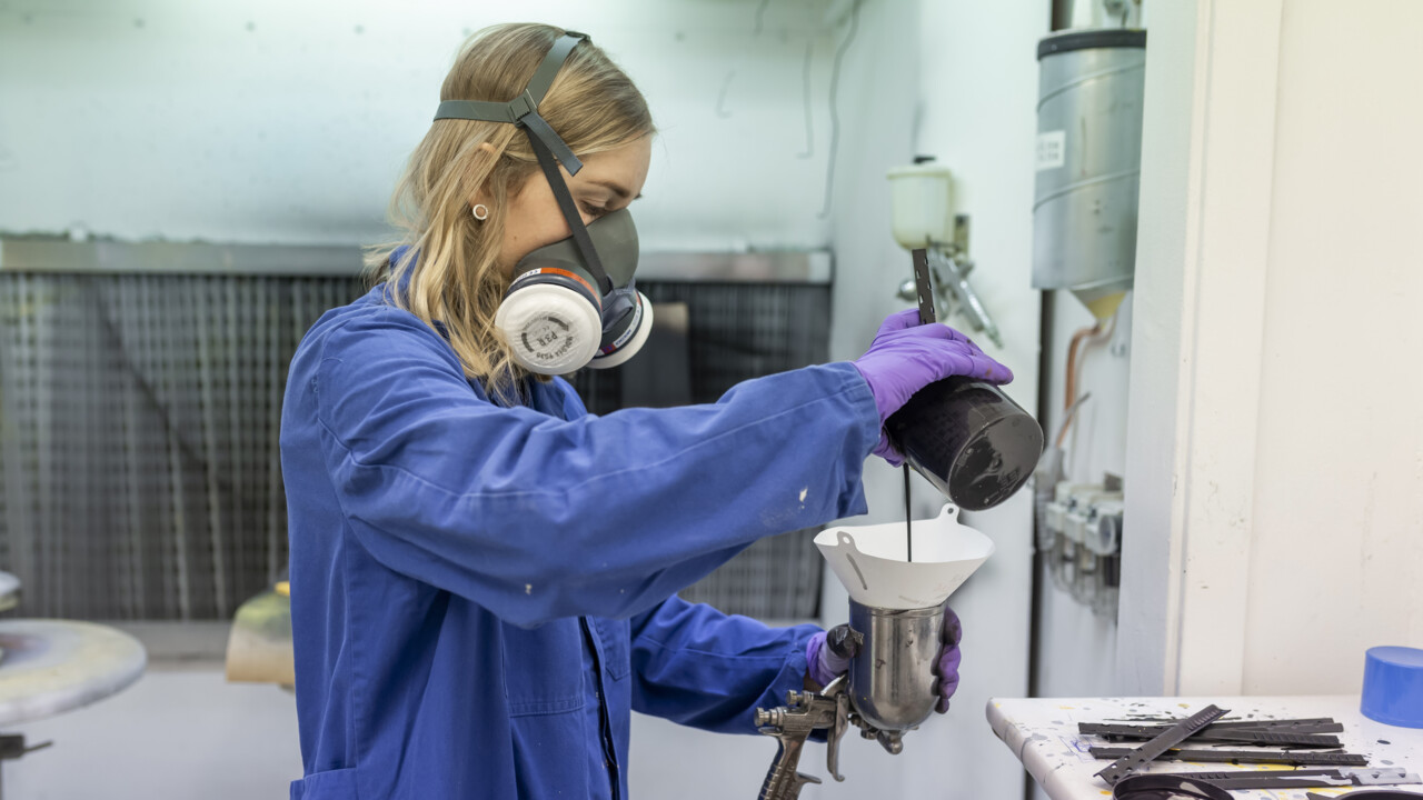 En ung kvinna som bär en mask över mun och näsa häller färg ur en glasbehållare ner i en färgspruta genom ett vitt filter. Hon bär en blå skyddsrock och lila gummihandskar.