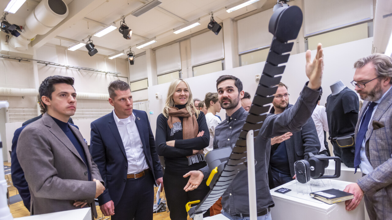 En ung man sträcker ut sina armar för att demonstrera längden på något. Framför honom är en prototyp. Fem personer står runt honom och lyssnar/tittar på det han demonstrerar. I bakgrunden syns många människor som befinner sig i samma lokal.